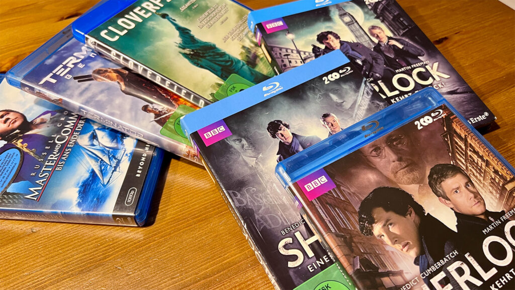 Auf dem Bild liegen verschiedene Blu-ray-Filme verstreut auf einem Tisch. Die Titel sind Master and Commander, Terminator Genysis, Cloverfield sowie die ersten drei Staffeln der Serie Sherlock.